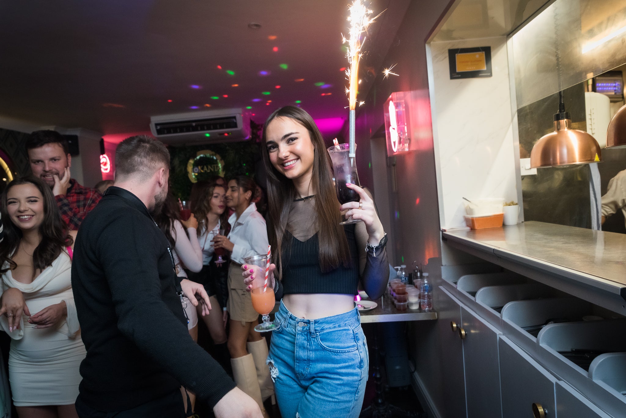 Woman holding a drink and sparkler in a lively bar setting with other people.