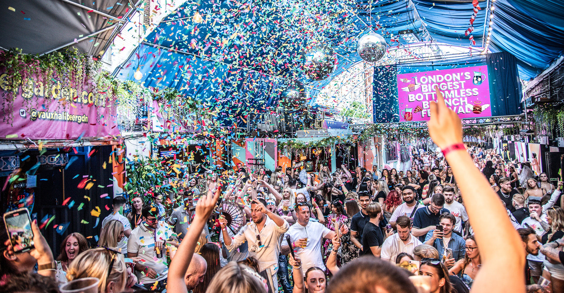 Crowd at a music festival with confetti, stage, and 'London's Biggest Bottomless' sign.