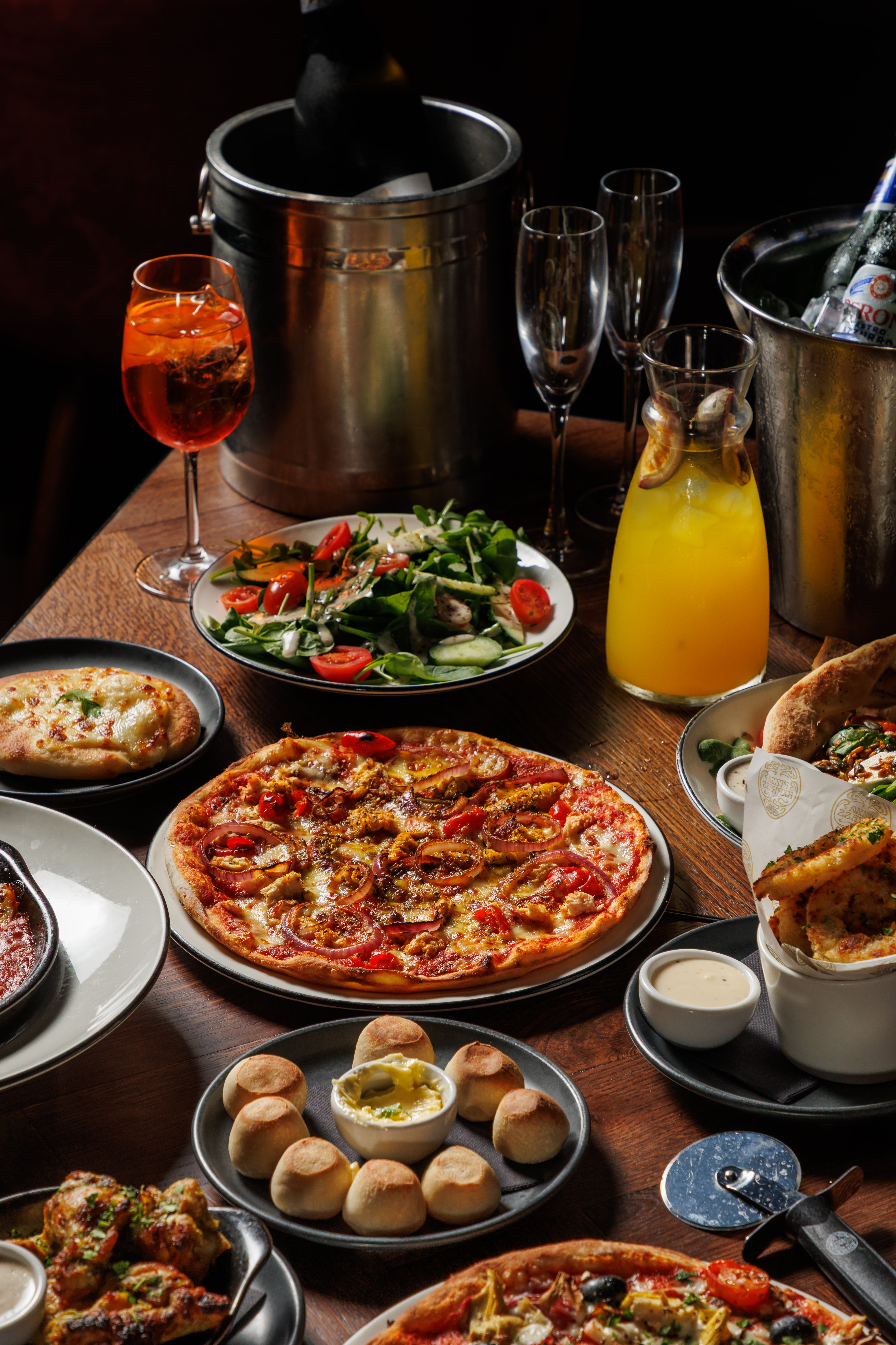 Table with various dishes including pizzas, bread, salad, and drinks in a restaurant setting.