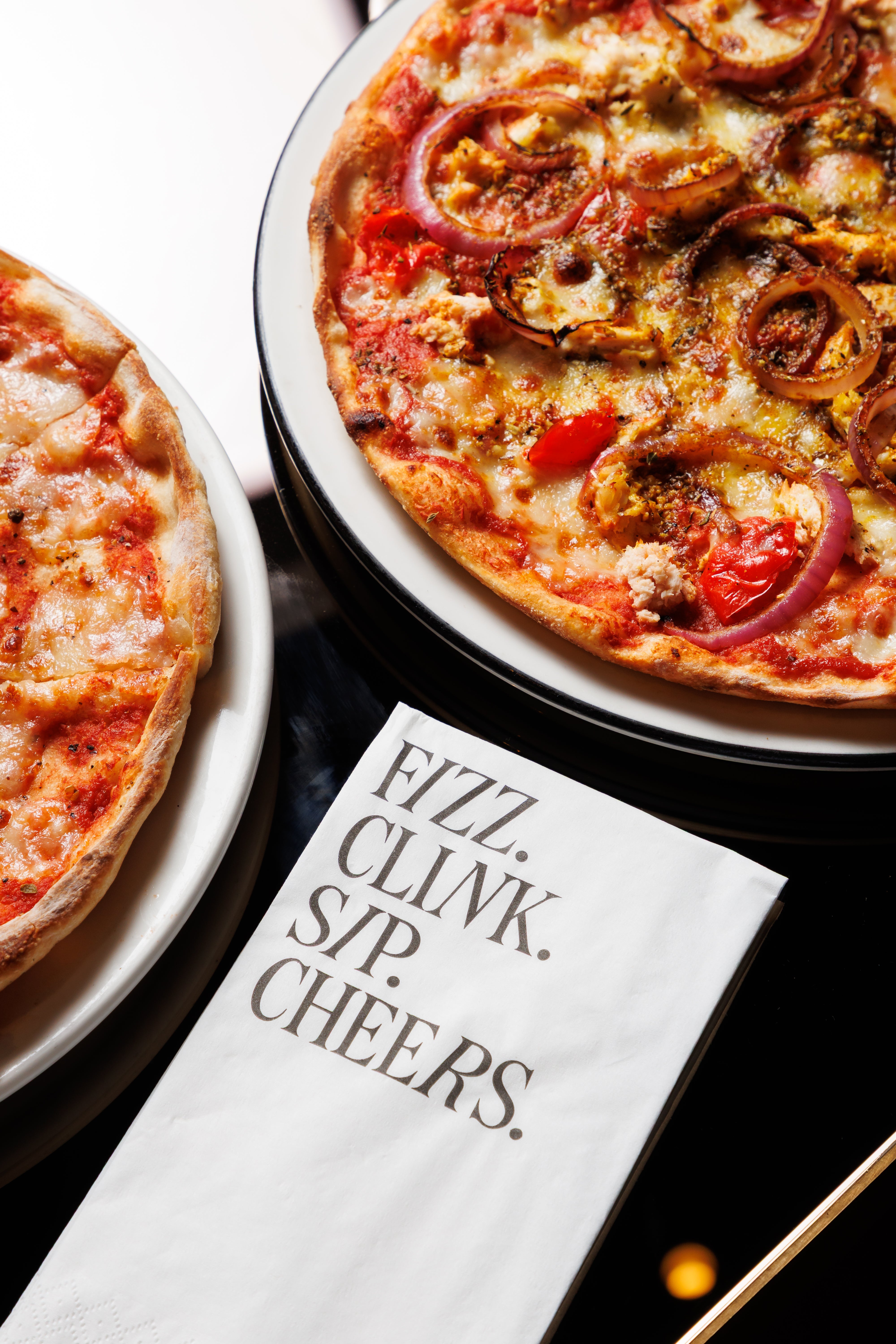 Two pizzas on plates with a napkin that says 'Fizz. Clink. Sip. Cheers.'