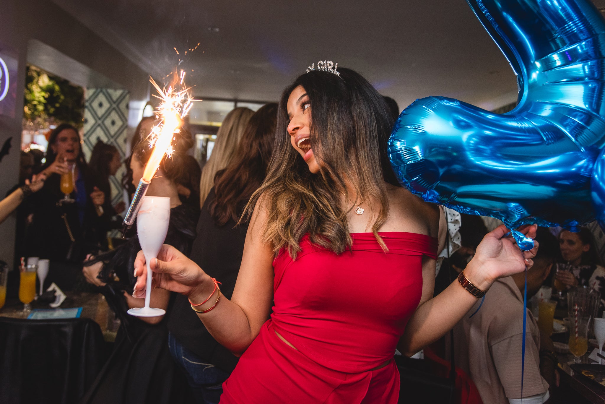 Woman in a red dress at a bottomless brunch holding a blue balloon and sparkler at a party for her birthday.